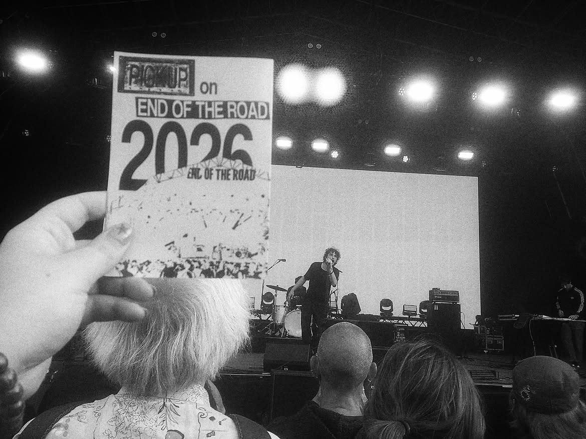 The PICKUP End of the Road 2026 zine held up in front of a band playing on stage